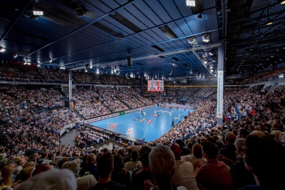 Eine große, mit Zuschauern gefüllte Halle, in der ein Handballspiel auf einem blauen Spielfeld unter hellen Lichtern und mit einer über dem Spielfeld hängenden Anzeigetafel läuft.