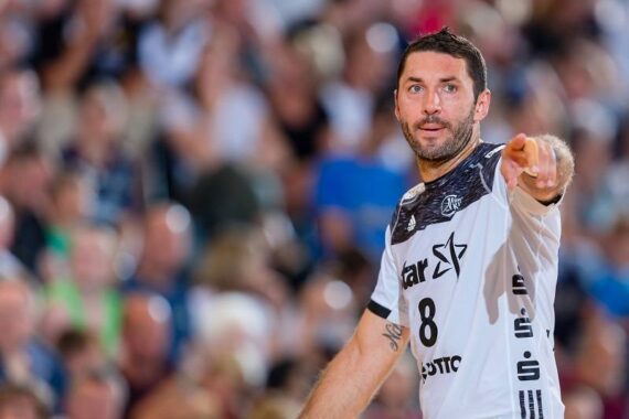 Ein männlicher Handballspieler in einem weiß-schwarzen Trikot mit der Nummer 8 zeigt nach vorne. Er befindet sich auf einem Spielfeld mit einer unscharfen Zuschauermenge im Hintergrund.