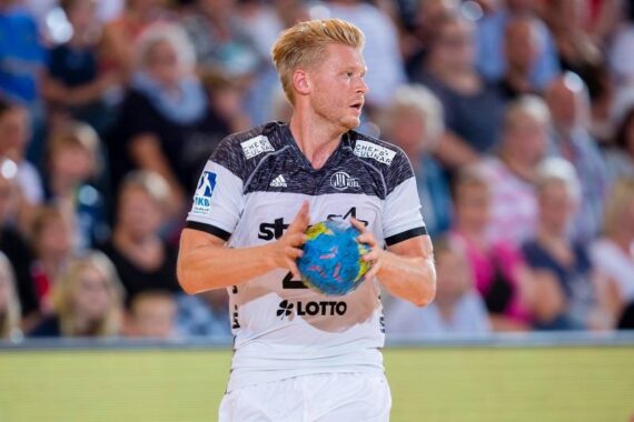 Ein männlicher Handballspieler in einem weiß-schwarzen Trikot hält einen blau-grünen Ball und blickt während eines Spiels nach rechts, mit einer unscharfen Menschenmenge im Hintergrund.