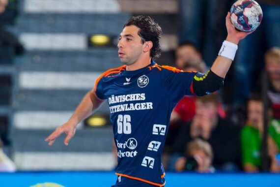 Ein männlicher Handballspieler in einem blau-orangenen Trikot mit der Nummer 66 bereitet sich während eines Spiels auf den Wurf vor. Im Hintergrund sind unscharfe Zuschauer und Stadionstufen zu sehen.