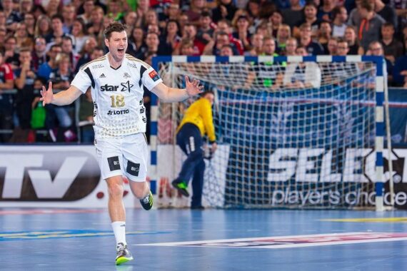 Ein männlicher Handballspieler in einem weißen Trikot jubelt mit ausgestreckten Armen auf einem Hallenplatz, während ein Torwart in Gelb in der Nähe des Tores steht und eine große Menschenmenge im Hintergrund zusieht.