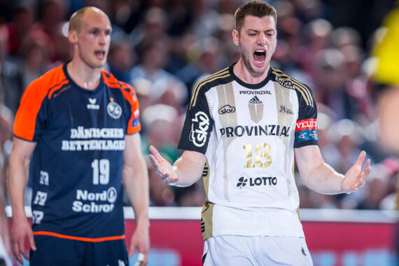 Ein männlicher Handballspieler in einem weiß-schwarzen Trikot ruft mit ausgestreckten Armen, während ein anderer Spieler in einem blau-orangenen Trikot in der Nähe steht. Sie befinden sich auf einem Hallenplatz mit einer unscharfen Menschenmenge im Hintergrund.