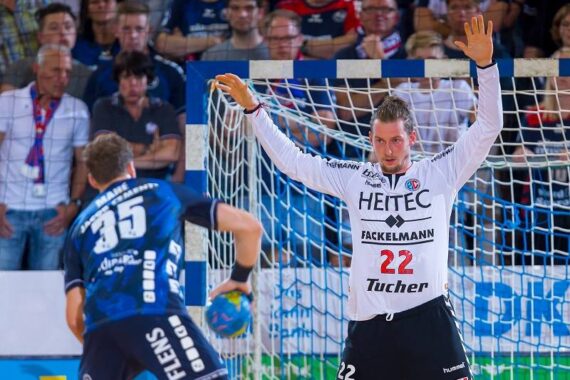 Ein Handballtorwart in einem weißen Trikot steht mit erhobenen Armen da und bereitet sich darauf vor, einen Schuss eines gegnerischen Spielers in einem blauen Trikot abzuwehren. Die Aktion findet in einer Halle statt, wo eine Menschenmenge hinter einem Netz zuschaut.