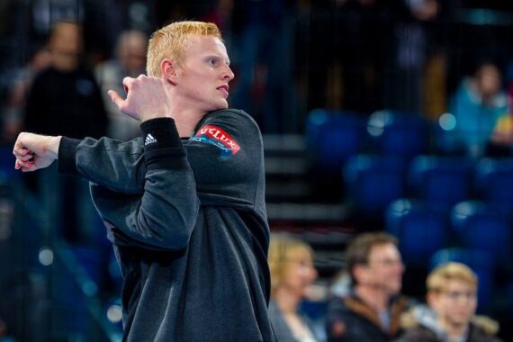 Ein Mann mit kurzen roten Haaren streckt seinen Arm in einer Sportarena über seine Brust und trägt eine schwarze Jacke. Im Hintergrund sind unscharfe Zuschauer und blaue Sitze zu sehen.