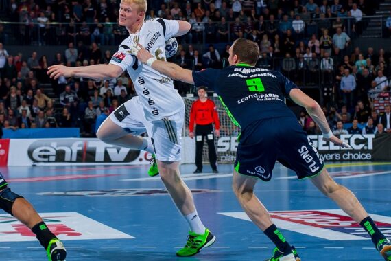Zwei männliche Handballspieler sind auf einem Hallenplatz in Aktion; ein Spieler in einer weißen Uniform springt, um den Ball zu werfen, während ein Verteidiger in einer marineblauen Uniform versucht, ihn zu blockieren. Die Zuschauer füllen die Tribünen im Hintergrund.