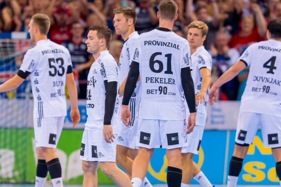 Eine Gruppe von männlichen Handballspielern in weißen Trikots mit schwarzen Akzenten steht auf einem Hallenplatz zusammen und bereitet sich auf das Spiel vor. Auf ihren Trikots sind Nummern und Sponsoren abgebildet. Unscharfe Menschenmenge im Hintergrund.