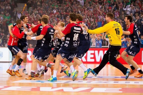 Eine Gruppe männlicher Handballspieler in roten, blauen und gelben Trikots feiert ausgelassen auf einem Hallenplatz, während im Hintergrund eine jubelnde Menge zu sehen ist.
