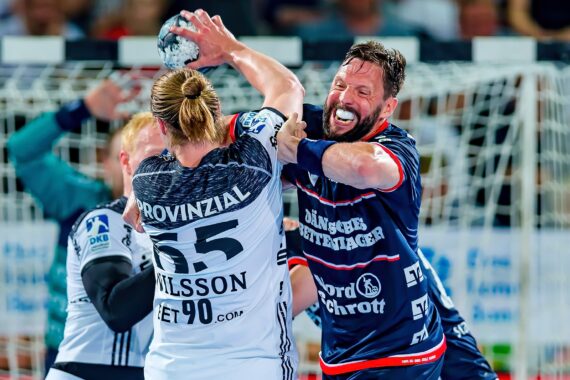 Zwei männliche Handballspieler in Aktion: Einer im weißen Trikot mit der Nummer 25 hält den Ball, während ein Spieler im blauen Trikot aggressiv verteidigt. Beide zeigen intensive Gesichtsausdrücke vor dem Tornetz.