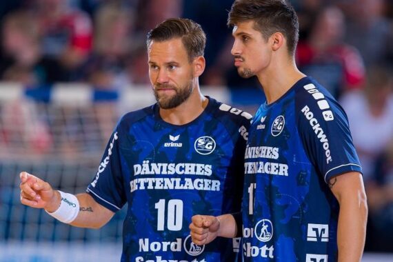 Zwei männliche Handballspieler in blauen Trikots mit Sponsorenlogos stehen in einer Halle nebeneinander und wirken während eines Spiels konzentriert und kommunikativ.