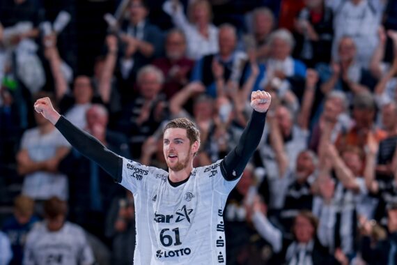 Ein Handballspieler in einem weißen Trikot hebt lächelnd beide Arme zum Jubel, während im Hintergrund eine jubelnde Menge steht. Das Publikum scheint begeistert und unterstützend zu sein.