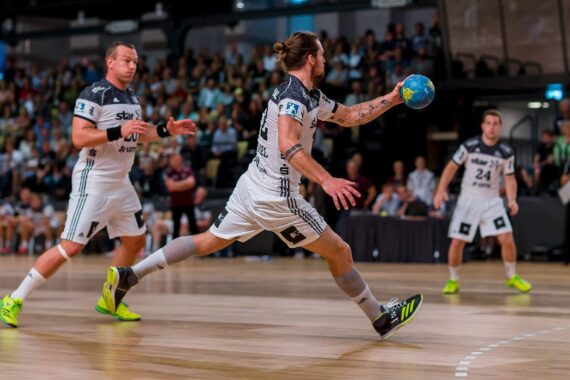 Ein Handballspieler in weißem Trikot springt nach vorne und bereitet sich darauf vor, einen blau-gelben Ball zu werfen, während seine Mannschaftskameraden und die Zuschauer in einer hell erleuchteten Halle zusehen.
