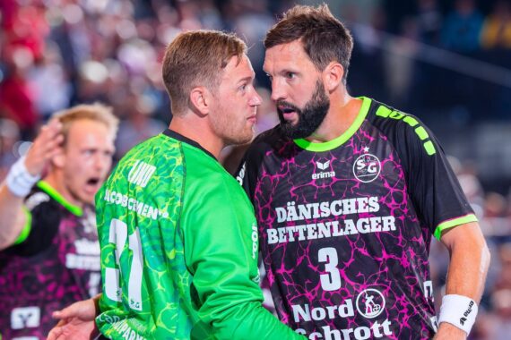 Zwei männliche Handballspieler im Gespräch auf dem Spielfeld, einer in einem grünen Trikot, der andere in einem schwarz-rosa Trikot, mit einer unscharfen Menschenmenge im Hintergrund.
