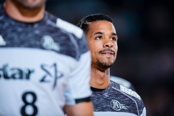 Ein männlicher Handballspieler in einem schwarz-weißen Trikot steht auf einem Spielfeld, blickt nach vorne und lächelt leicht. Ein anderer Spieler mit der Nummer 8 auf seinem Trikot ist teilweise im Vordergrund zu sehen.