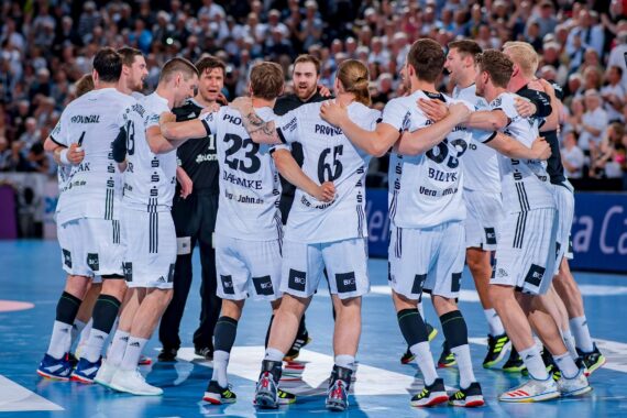 Eine Herren-Handballmannschaft in weißen Trikots steht in einem Kreis auf einem Hallenplatz, die Arme umeinander gelegt, und scheint zu feiern oder Strategien zu entwickeln, während im Hintergrund eine jubelnde Menge steht.