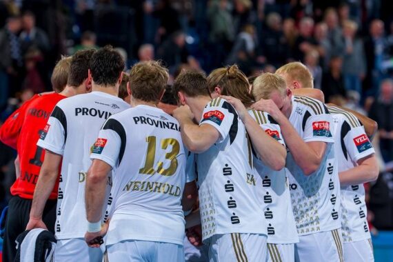 Eine Herren-Handballmannschaft in weißen Trikots kauert auf dem Spielfeld zusammen, die Arme umeinander gelegt, mit dem Rücken zur Kamera. Das Publikum ist unscharf im Hintergrund zu sehen.