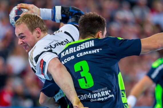 Zwei männliche Handballspieler in Aktion; der eine im weißen Trikot versucht zu werfen, während er von einem Spieler im blauen Trikot mit dem Namen Karlsson und der Nummer 3 eng verteidigt wird. Beide sind konzentriert, in einer überfüllten Halle.