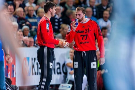 Zwei männliche Handballspieler in roten Trikots stehen während eines Spiels am Spielfeldrand; einer trinkt aus einer Dose, während der andere ein Handtuch hält. Die Zuschauer füllen den unscharfen Hintergrund.