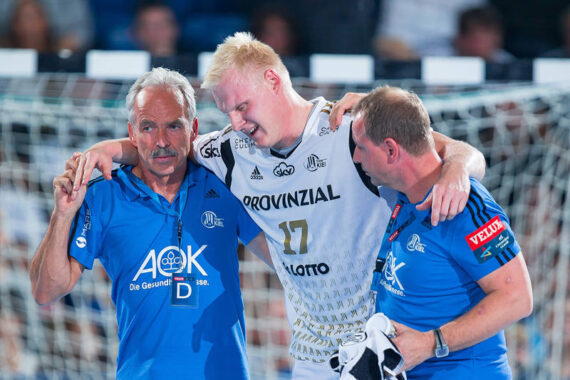Ein männlicher Sportler in einem weißen Trikot wird nach einer Verletzung während eines Handballspiels von zwei Mitarbeitern in blauen Hemden vom Spielfeld getragen, wobei im Hintergrund ein Tornetz zu sehen ist.