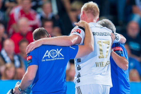 Ein Handballspieler in einem weißen Trikot, der von zwei Mitarbeitern in blauen Hemden unterstützt wird, wird vom Spielfeld getragen, was auf eine Verletzung während eines Spiels schließen lässt.