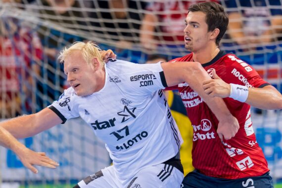 Zwei männliche Handballspieler treten während eines Spiels gegeneinander an; der Spieler in Weiß versucht, sich nach vorne zu bewegen, während der Spieler in Rot eng verteidigt; beide sind sehr konzentriert. Im Hintergrund sind ein Netz und unscharfe Zuschauer zu sehen.
