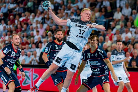 Ein Handballspieler in einem weißen Trikot springt zum Schuss, während er während eines Spiels von Verteidigern in dunkelblauen Trikots umringt ist, während die Zuschauer im Hintergrund zusehen.