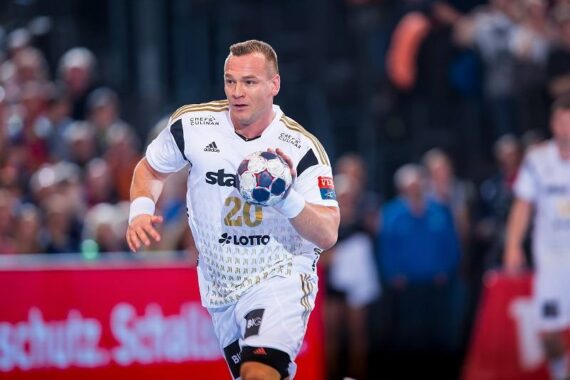 Ein männlicher Handballspieler in einem weißen Trikot mit der Nummer 20 läuft, während er während eines Spiels einen Ball hält. Der Hintergrund zeigt eine unscharfe Menge und andere Spieler.