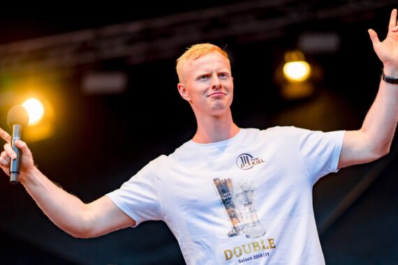 Ein Mann mit kurzen blonden Haaren hält ein Mikrofon in der Hand und hebt die andere Hand. Er steht auf der Bühne, hinter ihm sind helle Lichter zu sehen. Er trägt ein weißes T-Shirt mit einer Trophäengrafik und dem Wort DOUBLE darauf.