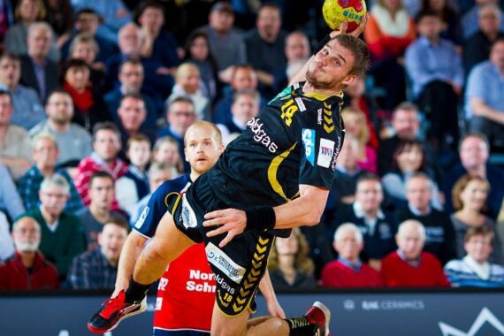 Ein männlicher Handballspieler in einem schwarz-gelben Trikot springt in der Luft und bereitet sich auf den Wurf vor, während ein Verteidiger in einem roten Trikot zusieht. Die Aktion findet vor einer Menge von Zuschauern in einer Halle statt.