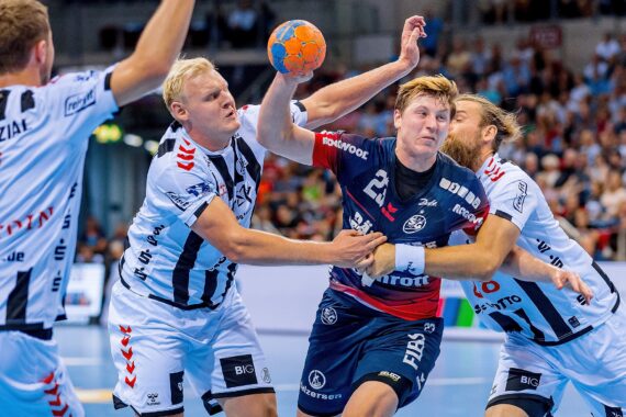 Drei Handballspieler in weißen Trikots verteidigen sich gegen einen Spieler in einem marineblauen Trikot, der springt und den Ball hält. Die Szene ist intensiv, mit Körperkontakt, während eines Spiels in einer überfüllten Halle.