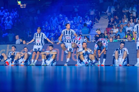 Eine Handballmannschaft in schwarz-weiß gestreiften Trikots sitzt und kniet auf dem Spielfeld und wirkt müde oder enttäuscht, während im Hintergrund eine Zuschauermenge bei blauer Beleuchtung zu sehen ist.