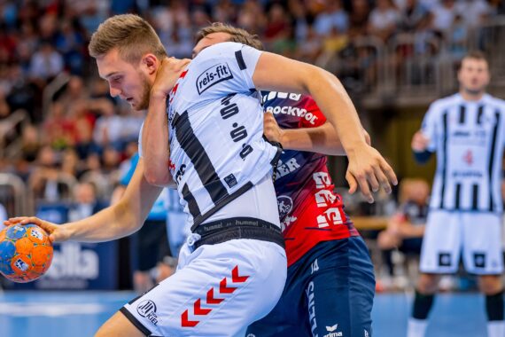 Ein Handballspieler in einem schwarz-weiß gestreiften Trikot wird während eines Spiels von einem gegnerischen Spieler in einem rot-blauen Trikot eng verteidigt und festgehalten, während im Hintergrund eine unscharfe Zuschauermenge zusieht.