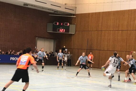 Ein Handballspiel in einer Turnhalle; der Spieler mit der Nummer 13 in Weiß springt zum Schuss, während die Verteidiger in Grau sich darauf vorbereiten, zu blocken. Der Spielstand ist 1:1 und die Zuschauer schauen von der Seite zu.