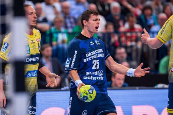 Ein männlicher Handballspieler in einem blauen Trikot (Nr. 25) schreit leidenschaftlich, während er den Ball hält, während zwei Spieler in gelben Trikots während eines Spiels mit einem Publikum im Hintergrund zusehen.