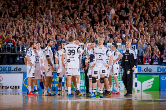 Eine Handballmannschaft in weißen Trikots versammelt sich auf dem Spielfeld, während im Hintergrund eine Gruppe von Zuschauern jubelt. Die Atmosphäre ist lebhaft, die Spieler und Trainer sind während einer scheinbaren Auszeit konzentriert.