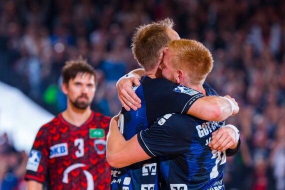 Zwei Handballspieler in blauen Trikots umarmen sich jubelnd, während ein Spieler in einem roten Trikot im Hintergrund steht und enttäuscht aussieht. Die Szene spielt sich in einer überfüllten Halle ab.