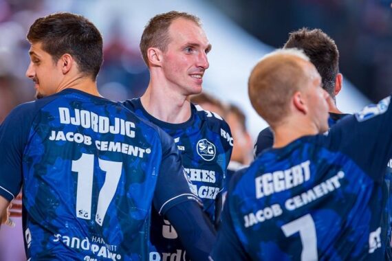 Drei männliche Handballspieler in blauen Trikots mit den Aufschriften DJORDJIC 17 und EGGERT 7 auf dem Rücken stehen zusammen auf einem Spielfeld, wobei ein Spieler lächelt und nach vorne schaut. Der Hintergrund ist unscharf.