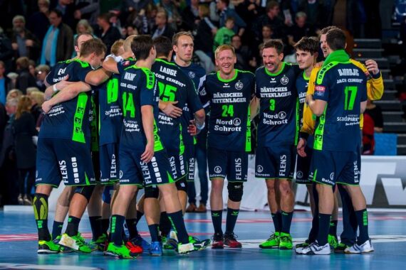 Eine Gruppe männlicher Handballspieler in marineblauen und neongrünen Uniformen steht nach einem Spiel lächelnd auf einem Hallenplatz und unterhält sich, während im Hintergrund Zuschauer zu sehen sind.