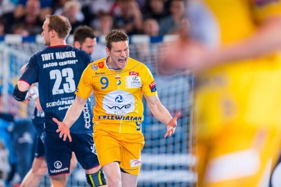 Ein männlicher Handballspieler im gelben Trikot mit der Nummer 9 schreit während eines Spiels leidenschaftlich mit ausgestreckten Armen. Ein anderer Spieler in einem dunkelblauen Trikot mit der Nummer 23 steht in der Nähe mit dem Rücken zur Kamera.