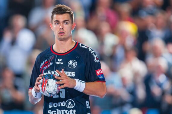 Ein männlicher Handballspieler in einem marineblauen Trikot hält einen Handball und schaut konzentriert. Im Hintergrund ist eine unscharfe Menschenmenge zu sehen, die auf eine Indoor-Sportveranstaltung hindeutet.