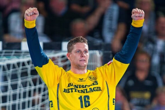Ein männlicher Handballspieler in einem gelb-blauen Trikot mit der Nummer 16 hebt vor einem Tor jubelnd beide Arme, im Hintergrund sind unscharfe Zuschauer zu sehen.