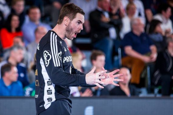 Ein männlicher Handballspieler in einem schwarzen Trikot gestikuliert heftig und schreit während eines Spiels, während im Hintergrund ein unscharfes Publikum zu sehen ist.