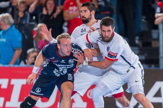 Drei männliche Handballspieler treten während eines Spiels gegeneinander an. Ein Spieler in einer marineblauen Uniform versucht, an zwei Verteidigern in weißen Uniformen vorbeizukommen, während im Hintergrund eine Menschenmenge zusieht.