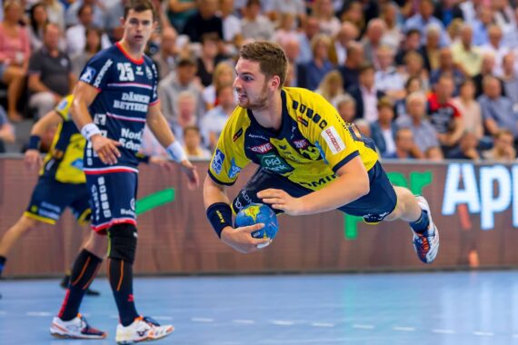 Ein Handballspieler in einem gelben Trikot springt mit einem Ball in der Hand durch die Luft und zielt auf ein Tor. Ein anderer Spieler in einem dunklen Trikot schaut zu, und im Hintergrund ist eine Menschenmenge zu sehen.