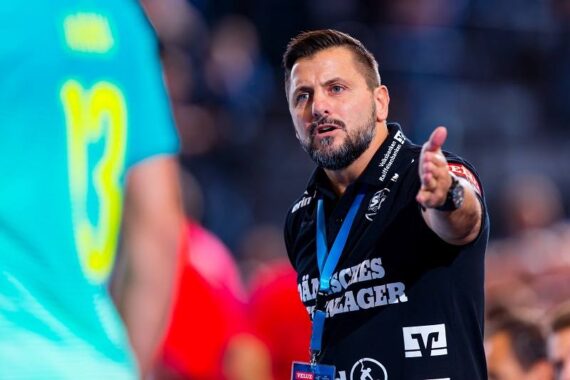 Ein Mann in einem schwarzen Hemd gestikuliert mit seiner rechten Hand, während er mit einer Person in einem türkisfarbenen Hemd mit der Nummer 13 spricht, während einer Hallensportveranstaltung. Der Hintergrund ist unscharf, andere Personen sind sichtbar.