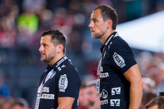 Zwei Männer in passenden schwarzen Hemden mit Sponsorenlogos stehen nebeneinander in einer Sportarena. Sie wirken konzentriert und aufmerksam und beobachten etwas im Hintergrund. Der Hintergrund ist unscharf, eine Menschenmenge ist zu sehen.