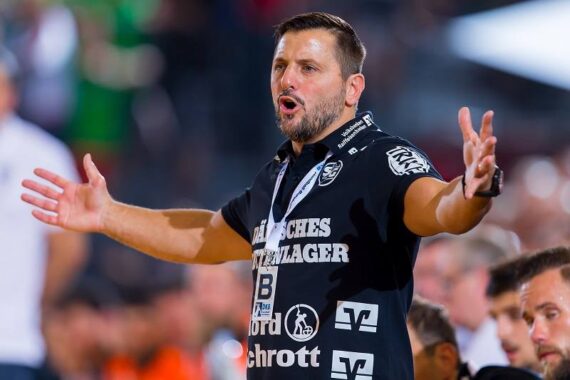 Ein Mann, der ein schwarzes Sporthemd trägt, gestikuliert mit weit ausgebreiteten Armen und scheint während einer Sportveranstaltung zu schreien, mit verschwommenen Menschen und hellen Lichtern im Hintergrund.