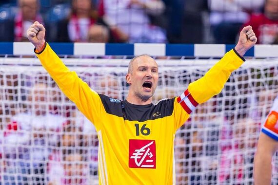Ein Handballtorwart in einem gelben Trikot mit der Nummer 16 feiert mit erhobenen Armen vor dem Tor, während die Zuschauer im Hintergrund von der Tribüne aus zusehen.