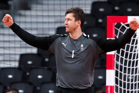 Ein männlicher Handballspieler in einer grau-schwarzen Uniform steht mit erhobenen Armen in einer Sportarena vor einem Tornetz.