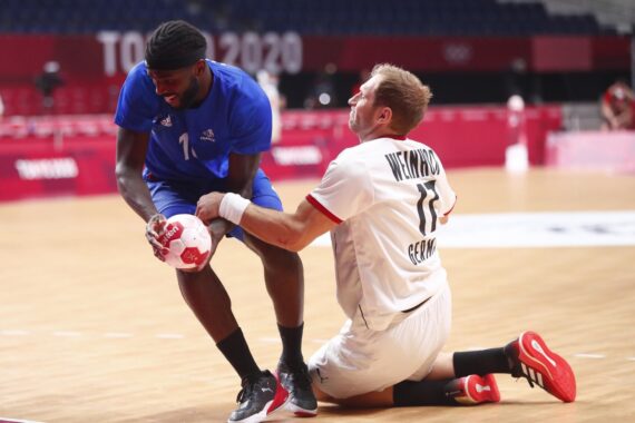 Ein französischer Handballspieler in Blau hält den Ball, während ein deutscher Spieler in Weiß, der auf dem Spielfeld kniet, während eines Spiels bei den Olympischen Spielen 2020 in Tokio zu verteidigen versucht.
