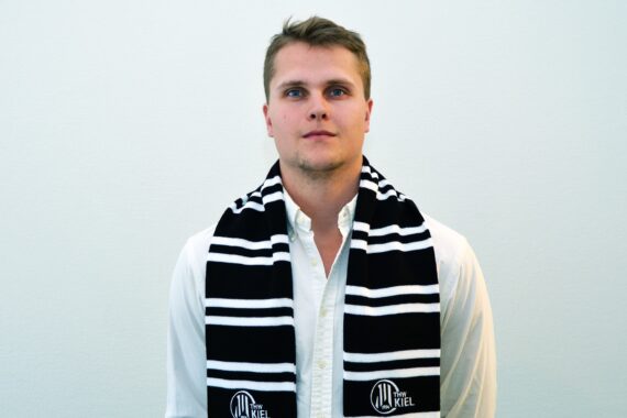Ein junger Mann mit weißem Hemd und schwarz-weiß gestreiftem Schal mit THW-Logo steht vor einer schlichten, hellen Wand und schaut mit neutralem Blick direkt in die Kamera.